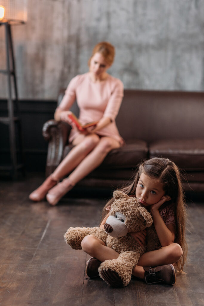 Young girl sitting alone with a teddy bear while an adult sits in the background, representing childhood trauma and emotional isolation.